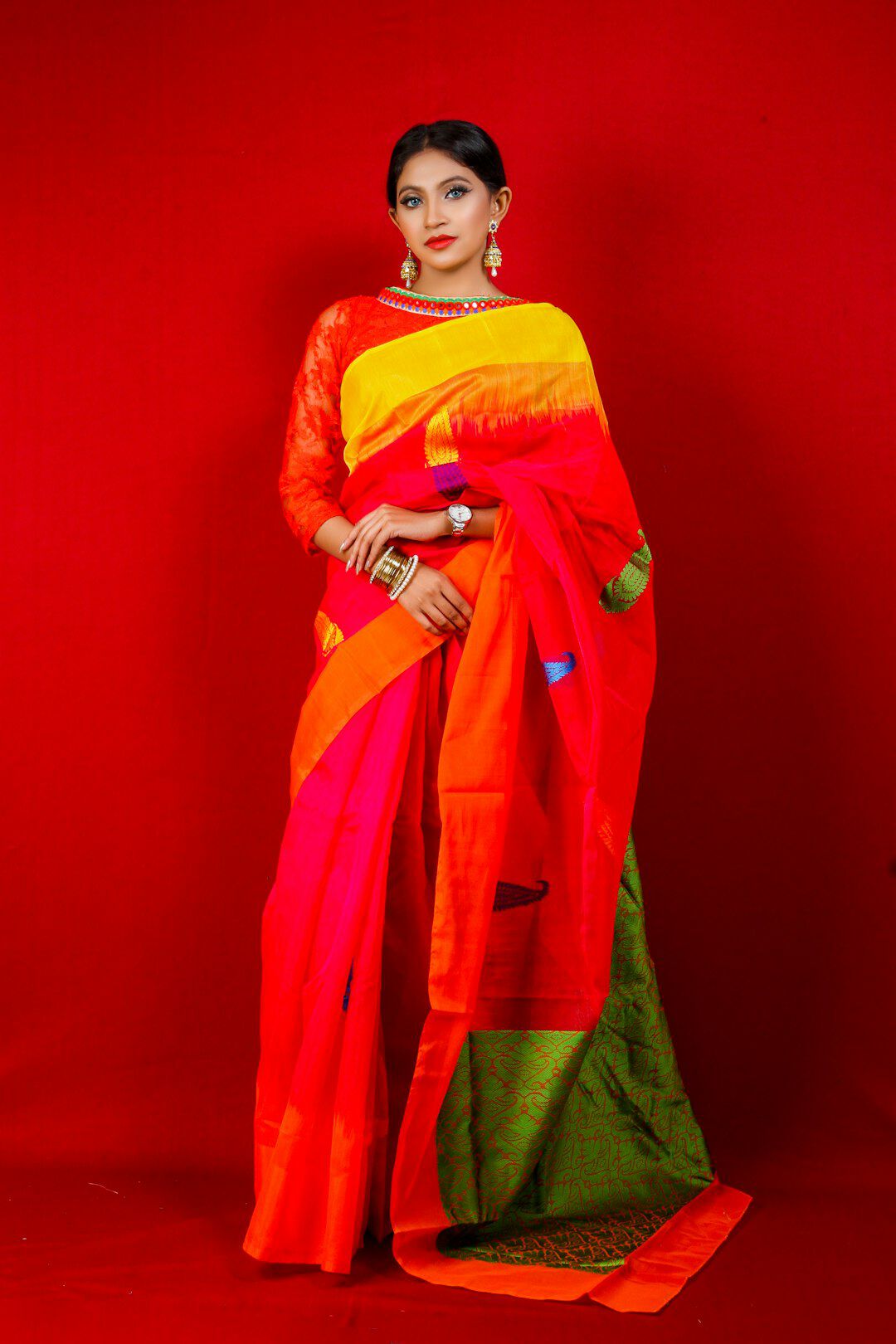Woman wearing a vibrant red, orange, and yellow silk saree with a green border, paired with traditional jewelry, posing against a bright red background